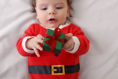 Cute baby wearing festive Christmas costume with gift box on white bedsheet, top view