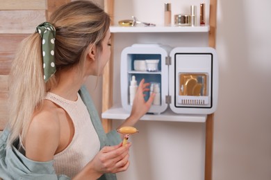 Photo of Woman with face roller taking cosmetic product from mini fridge indoors