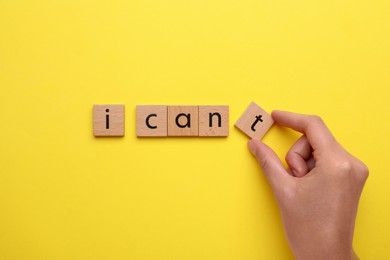 Motivation concept. Woman changing phrase from I Can't into I Can by removing wooden square with letter T on yellow background, top view