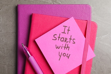 Photo of Note with phrase It Starts With You, pen and notebooks on grey table, top view