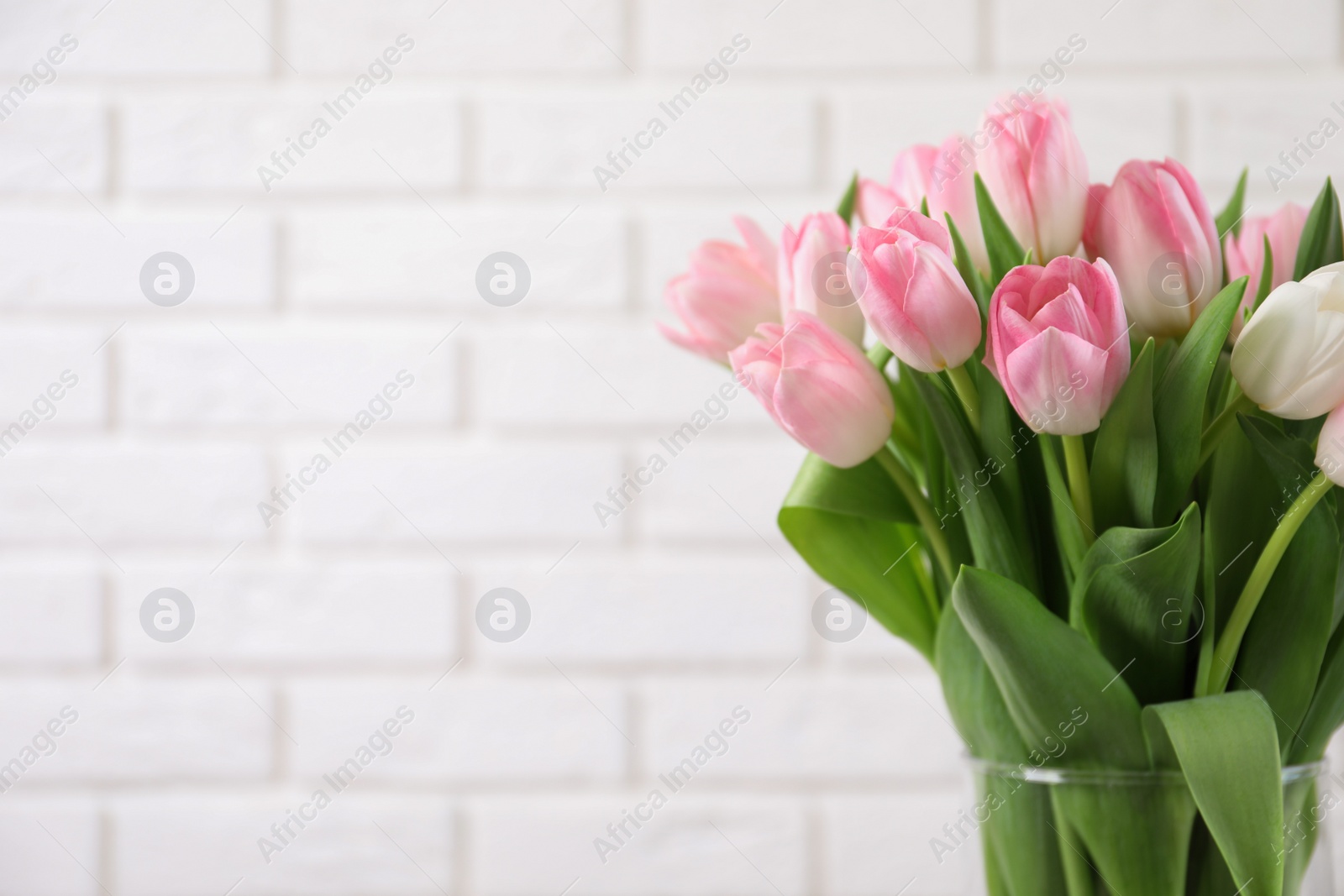 Photo of Beautiful bouquet of tulips in glass vase against white brick wall. Space for text