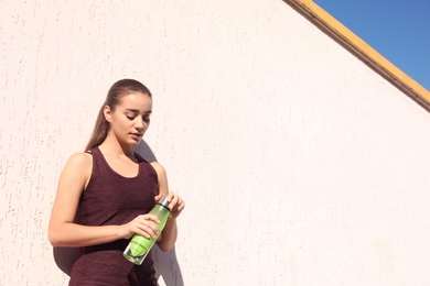 Photo of Young sporty woman holding bottle of water near wall outdoors on sunny day. Space for text