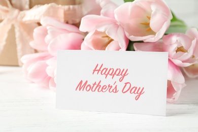 Photo of Greeting card with text HAPPY MOTHER'S DAY, tulips and gift box on table