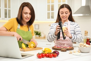 Photo of Mother with adult daughter making dinner while watching online cooking course via laptop in kitchen