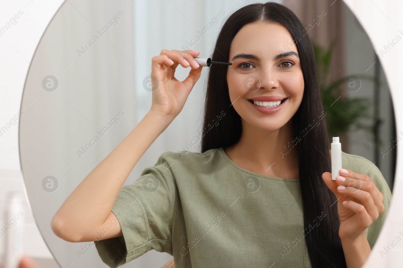 Photo of Beautiful woman applying serum onto her eyelashes in room, reflection in mirror. Cosmetic product