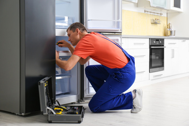 Male technician with screwdriver repairing refrigerator in kitchen