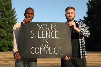 Photo of Men holding sign with phrase Your Silence Is Complicit outdoors. Racism concept