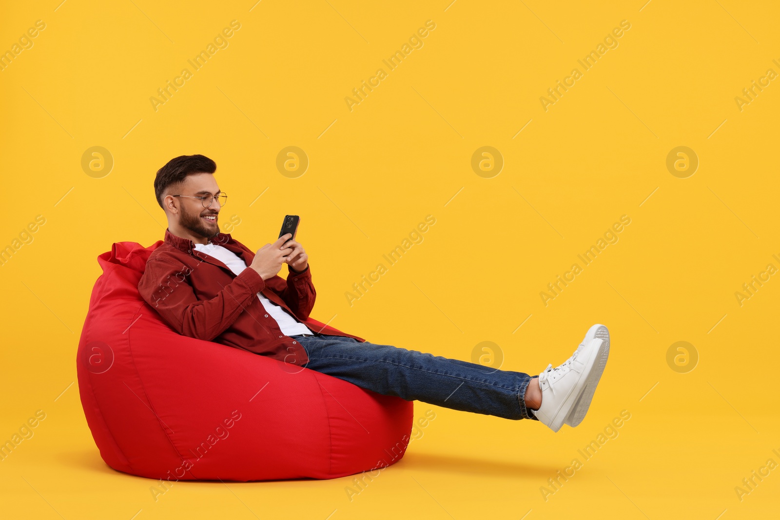 Photo of Happy young man using smartphone on bean bag chair against yellow background, space for text