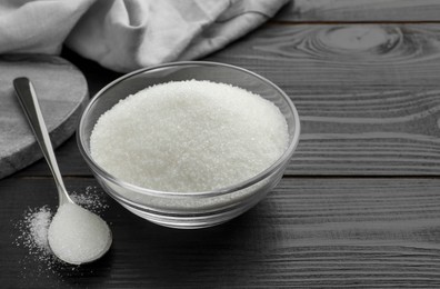 Photo of Granulated sugar in bowl and spoon on black wooden table, closeup. Space for text