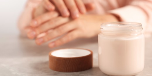 Image of Woman applying cosmetic cream onto hand at grey table, focus on jar. Banner design