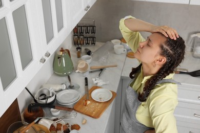 Photo of Stressed young woman in messy kitchen. Many dirty dishware, utensils and food leftovers on white countertop