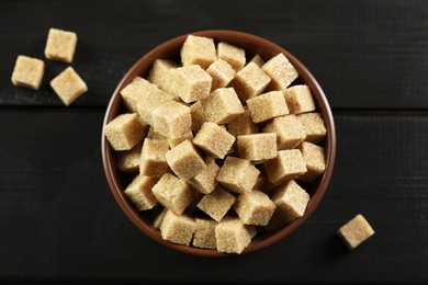 Photo of Brown sugar cubes in bowl on black wooden table, top view