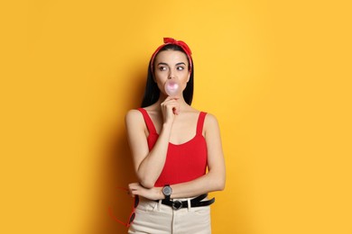 Photo of Fashionable young woman in pin up outfit blowing bubblegum on yellow background