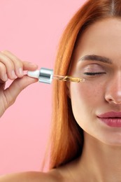 Beautiful young woman applying cosmetic serum onto her face on pink background, closeup