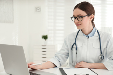Photo of Young female doctor working with laptop at table in office