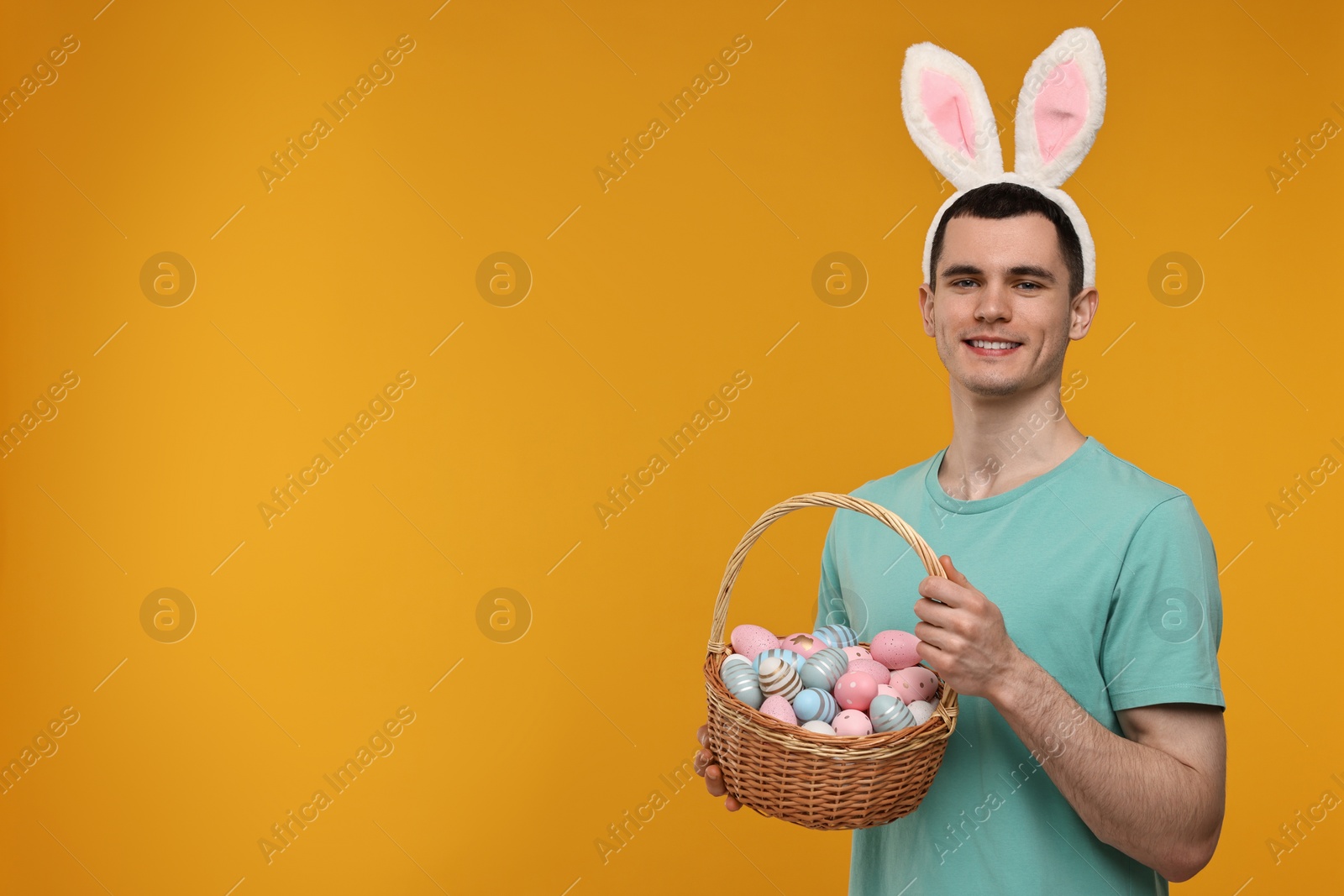 Photo of Easter celebration. Handsome young man with bunny ears holding basket of painted eggs on orange background. Space for text