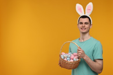 Photo of Easter celebration. Handsome young man with bunny ears holding basket of painted eggs on orange background. Space for text