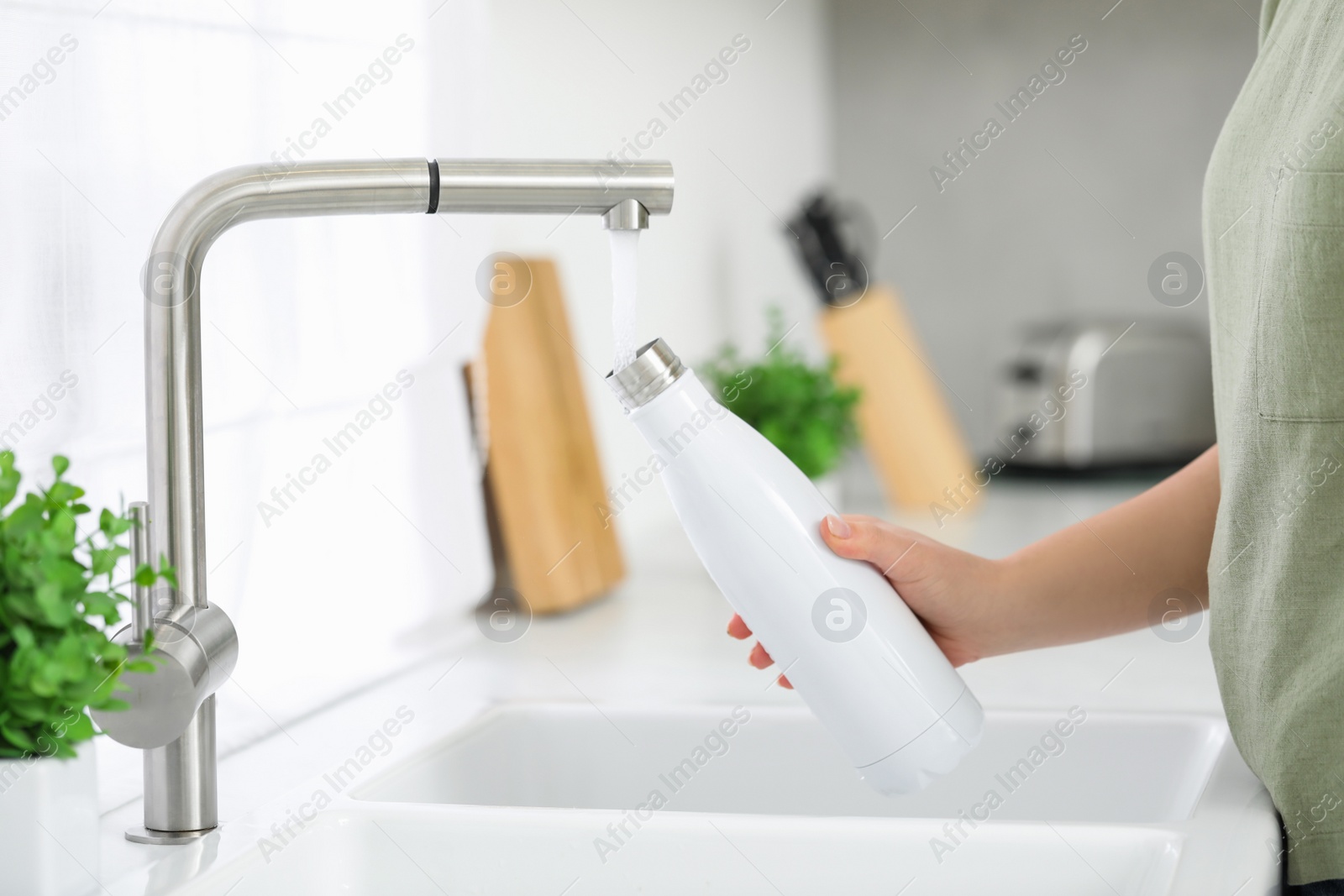 Photo of Woman pouring fresh water from tap into thermo bottle in kitchen, closeup