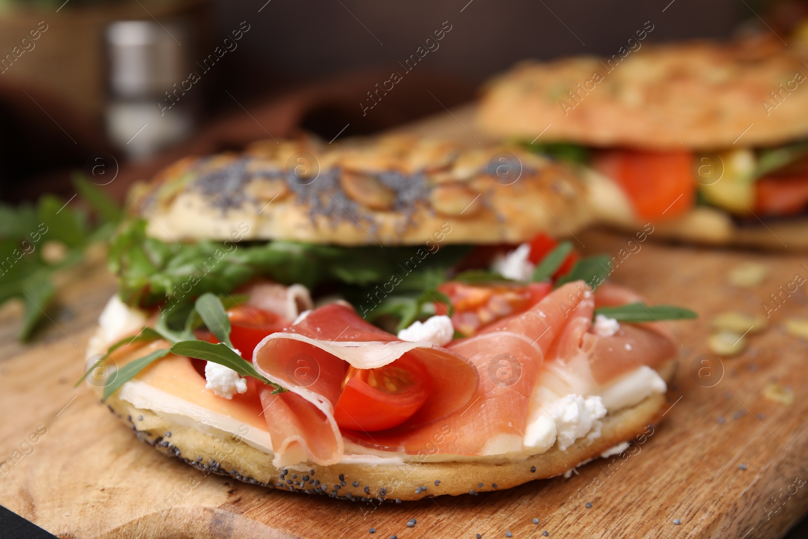 Photo of Tasty bagel with cured ham, cream cheese, tomatoes and arugula on wooden board, closeup