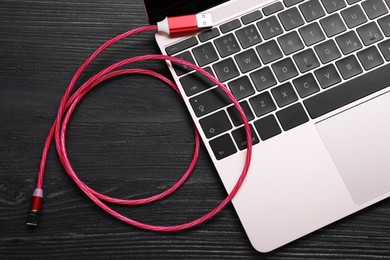 Red USB cable with type C connector and laptop on black wooden table, flat lay