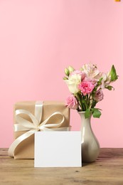 Happy Mother's Day. Gift box, blank card and bouquet of beautiful flowers in vase on wooden table against pink background