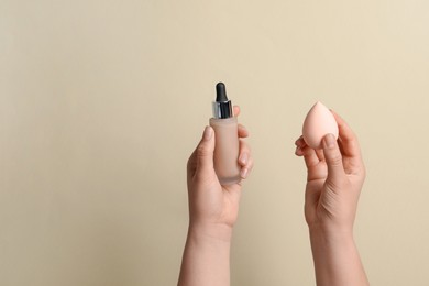 Photo of Woman holding bottle with skin foundation and makeup sponge on beige background, closeup. Space for text