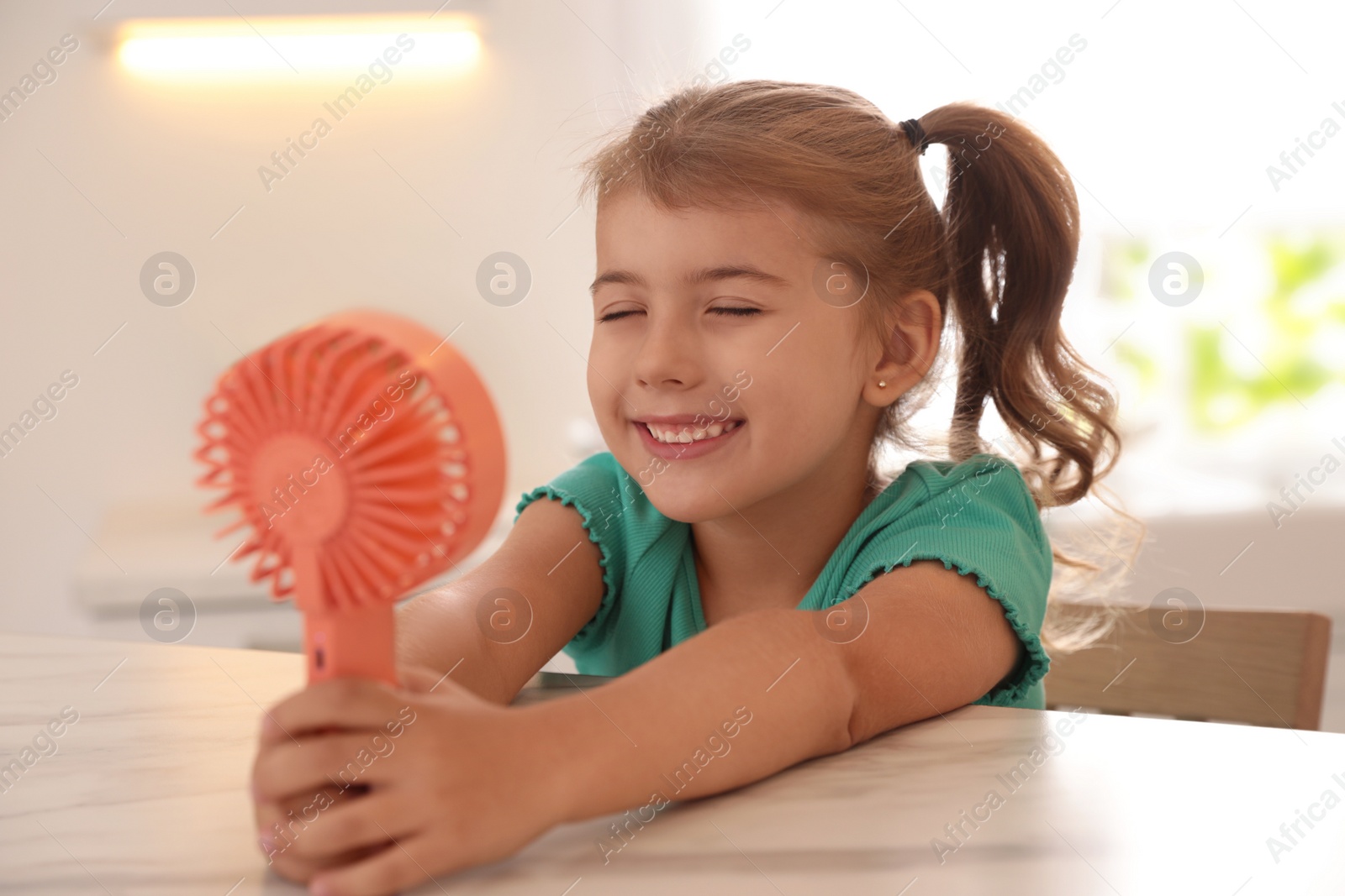 Photo of Little girl enjoying air flow from portable fan at table in kitchen. Summer heat