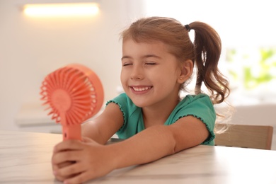 Little girl enjoying air flow from portable fan at table in kitchen. Summer heat