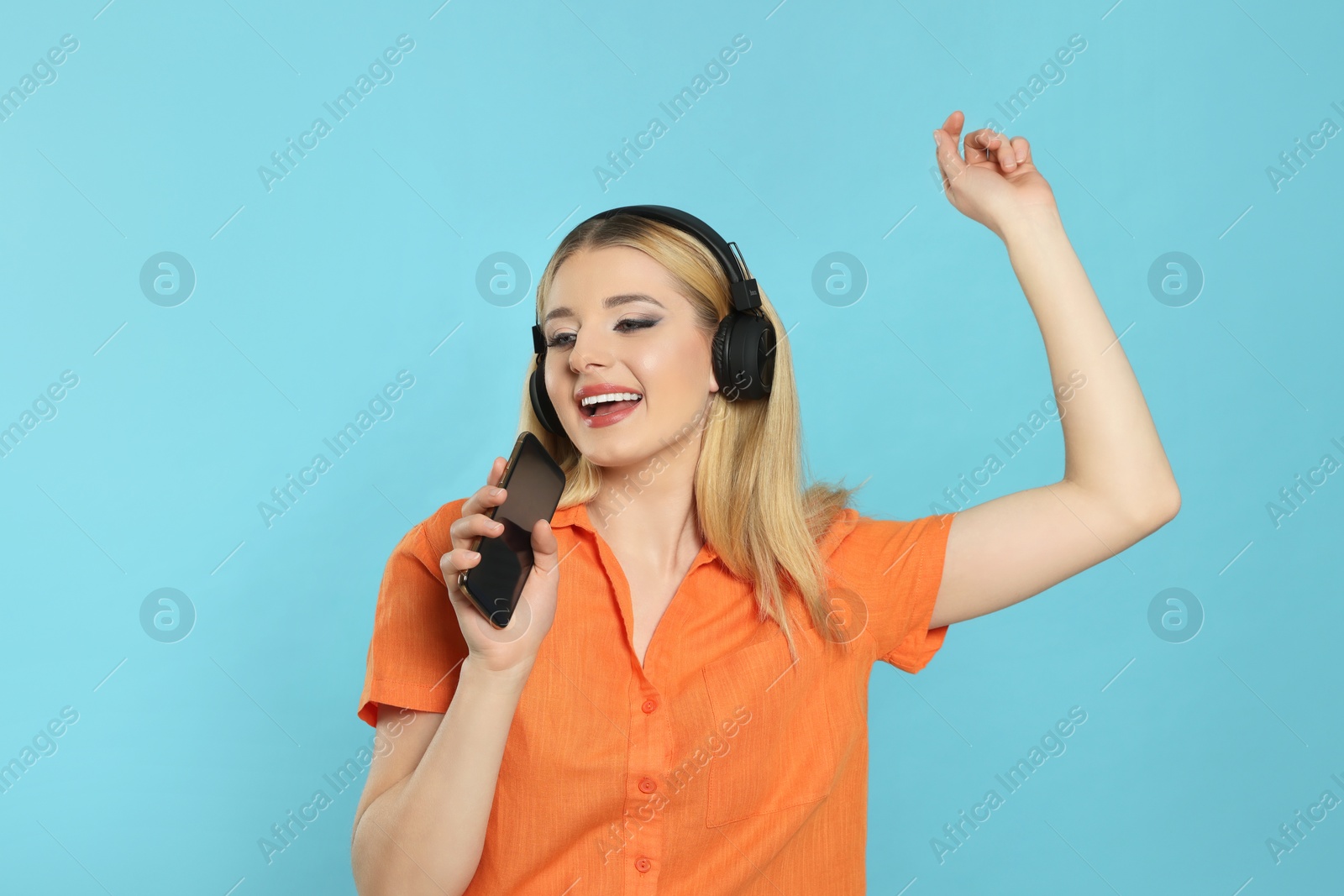 Photo of Happy woman in headphones enjoying music and singing into smartphone on light blue background