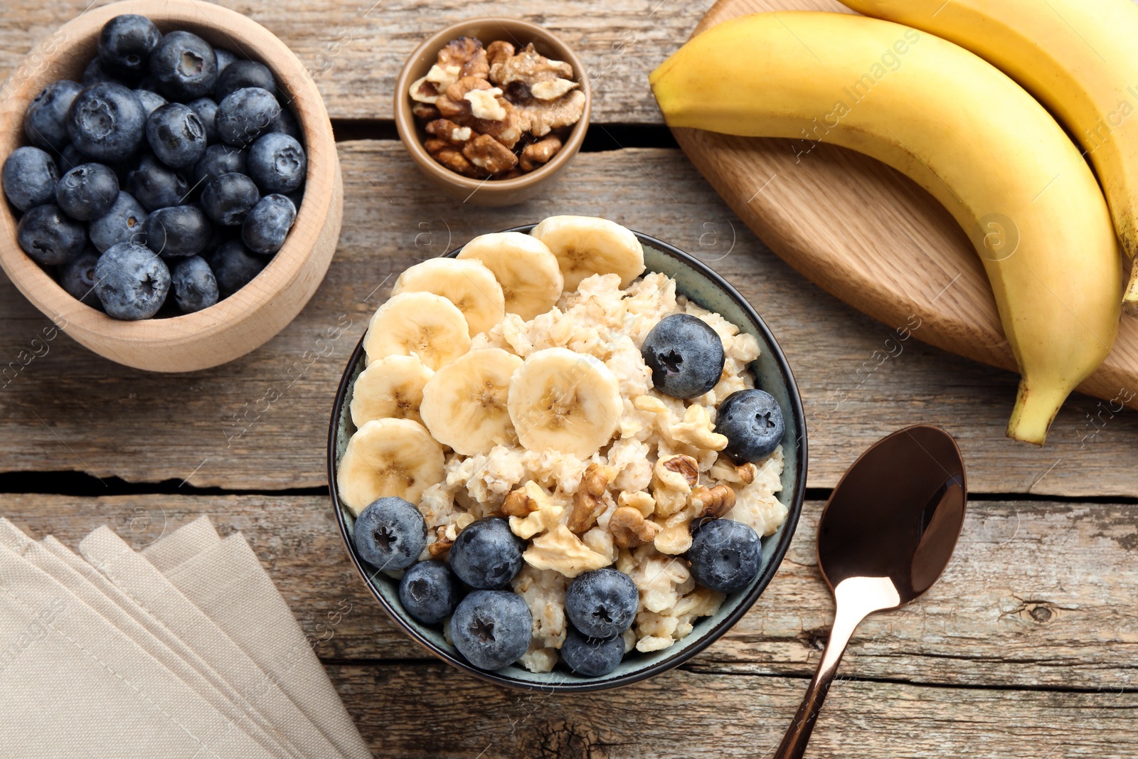 Photo of Tasty oatmeal with banana, blueberries and walnuts served in bowl on wooden table, flat lay