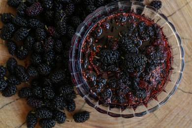 Photo of Delicious ripe black mulberries and bowl of sweet jam on wood stump, flat lay