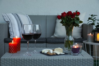 Glasses of wine, vase with roses, burning candles and snacks on rattan table at balcony in evening