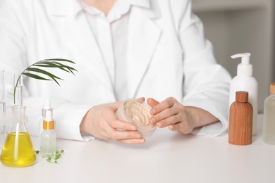 Photo of Dermatologist with jar testing cosmetic product at white table indoors, selective focus