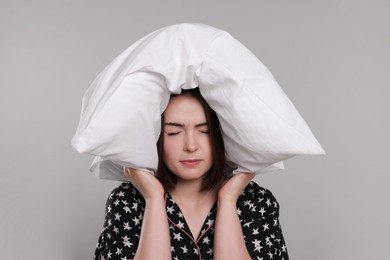 Unhappy young woman covering ears with pillow on light grey background. Insomnia problem