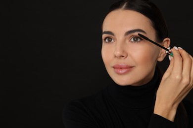 Beautiful young woman applying mascara on black background, closeup. Space for text