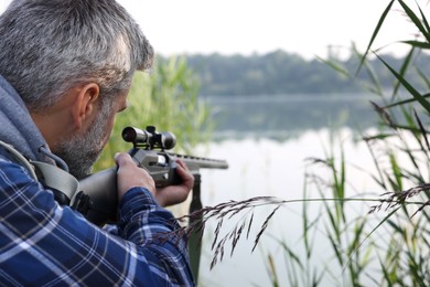 Photo of Man aiming with hunting rifle near lake outdoors. Space for text