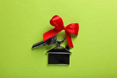 Key with trinket in shape of house and red bow on light green background, top view. Housewarming party