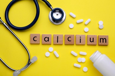 Photo of Word Calcium made of wooden cubes with letters, stethoscope and pills on yellow background, flat lay