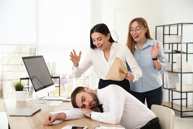 Photo of Young women popping paper bag behind their sleeping colleague in office. Funny joke
