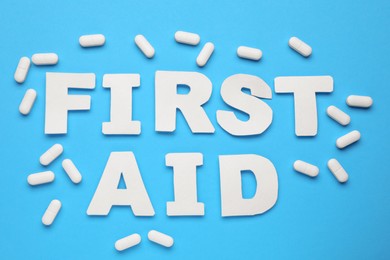 Photo of Words First Aid made of white letters and pills on light blue background, flat lay