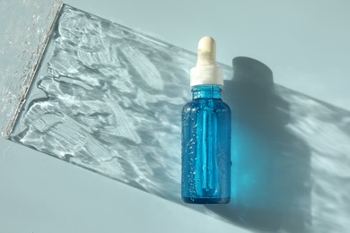 Wet bottle of cosmetic serum on light blue background, top view
