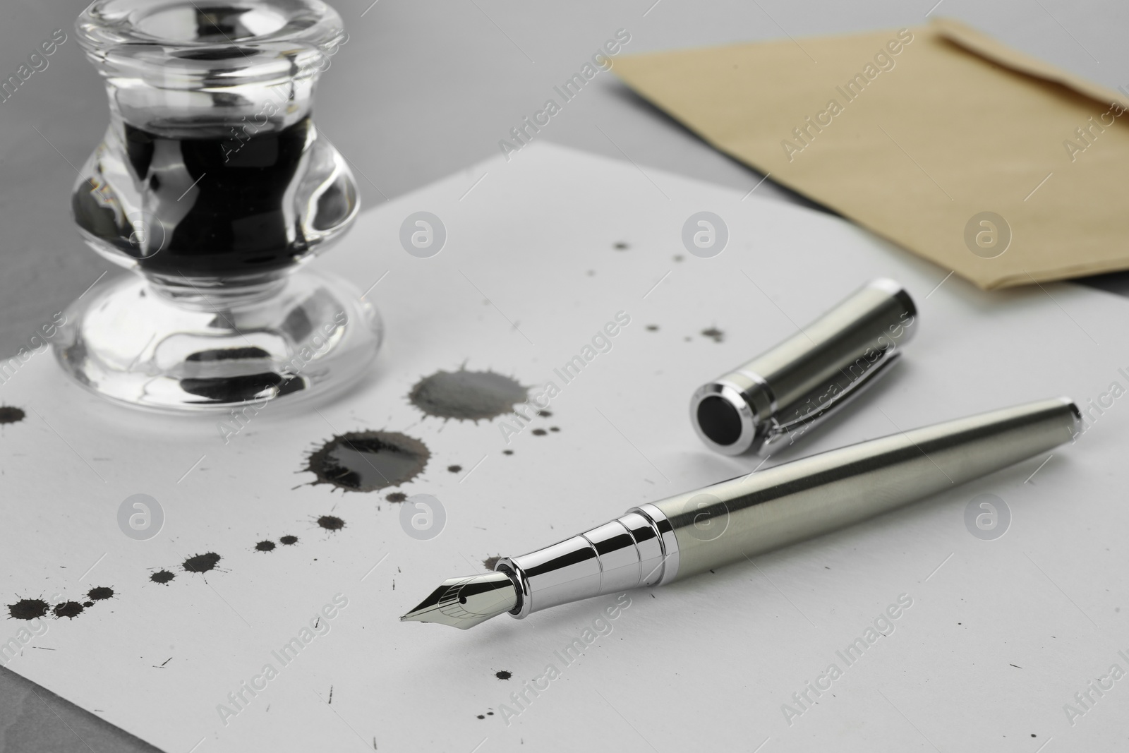 Photo of Stylish fountain pen, paper with blots of ink and inkwell on light grey table