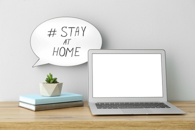 Photo of Laptop, books, houseplant and speech bubble with hashtag STAY AT HOME on white wall. Message to promote self-isolation during COVID‑19 pandemic