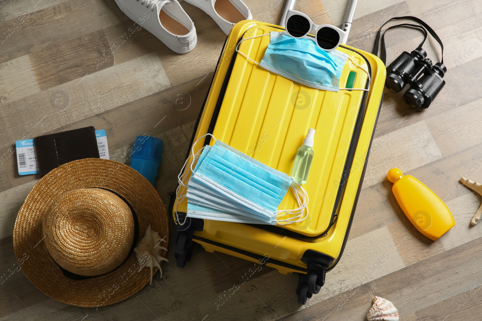 Photo of Flat lay composition with suitcase, protective masks, antiseptic spray and personal items on wooden floor. Travelling during coronavirus pandemic