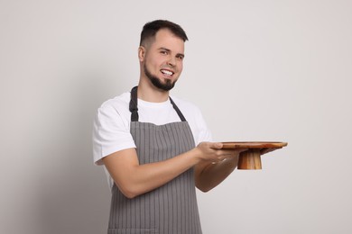 Photo of Happy professional confectioner in apron holding empty cake stand on light grey background