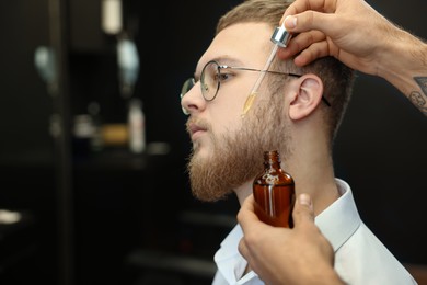 Photo of Hairdresser applying oil onto client's beard in barbershop, closeup. Professional shaving service