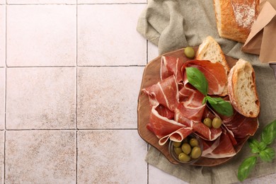 Slices of tasty cured ham, olives, bread and basil on tiled table, flat lay. Space for text