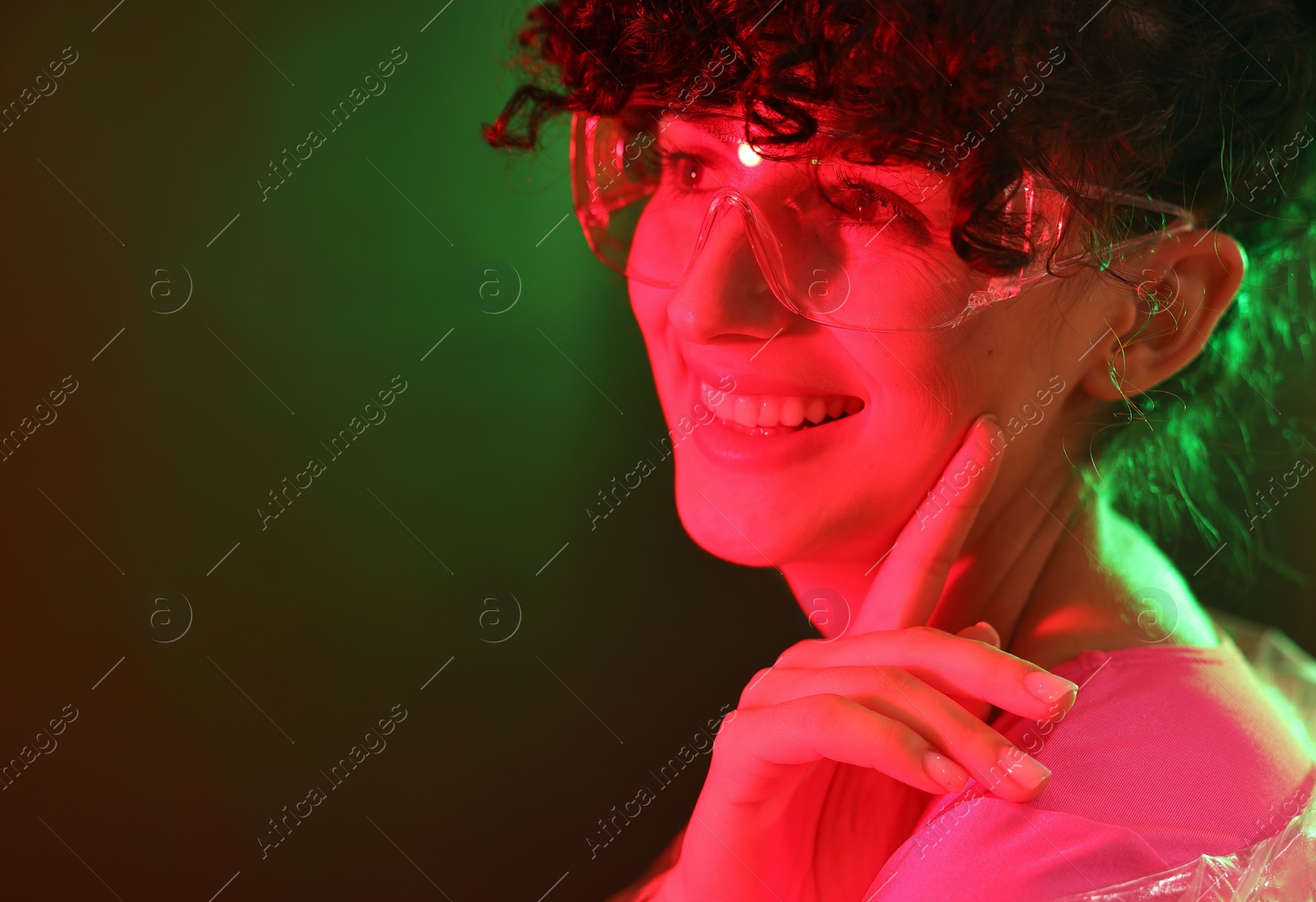 Photo of Beautiful young woman in transparent coat and sunglasses posing on color background in neon lights. Space for text