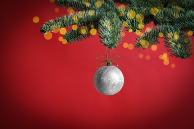 Photo of Beautiful Christmas ball hanging on fir tree branch against red background, bokeh effect