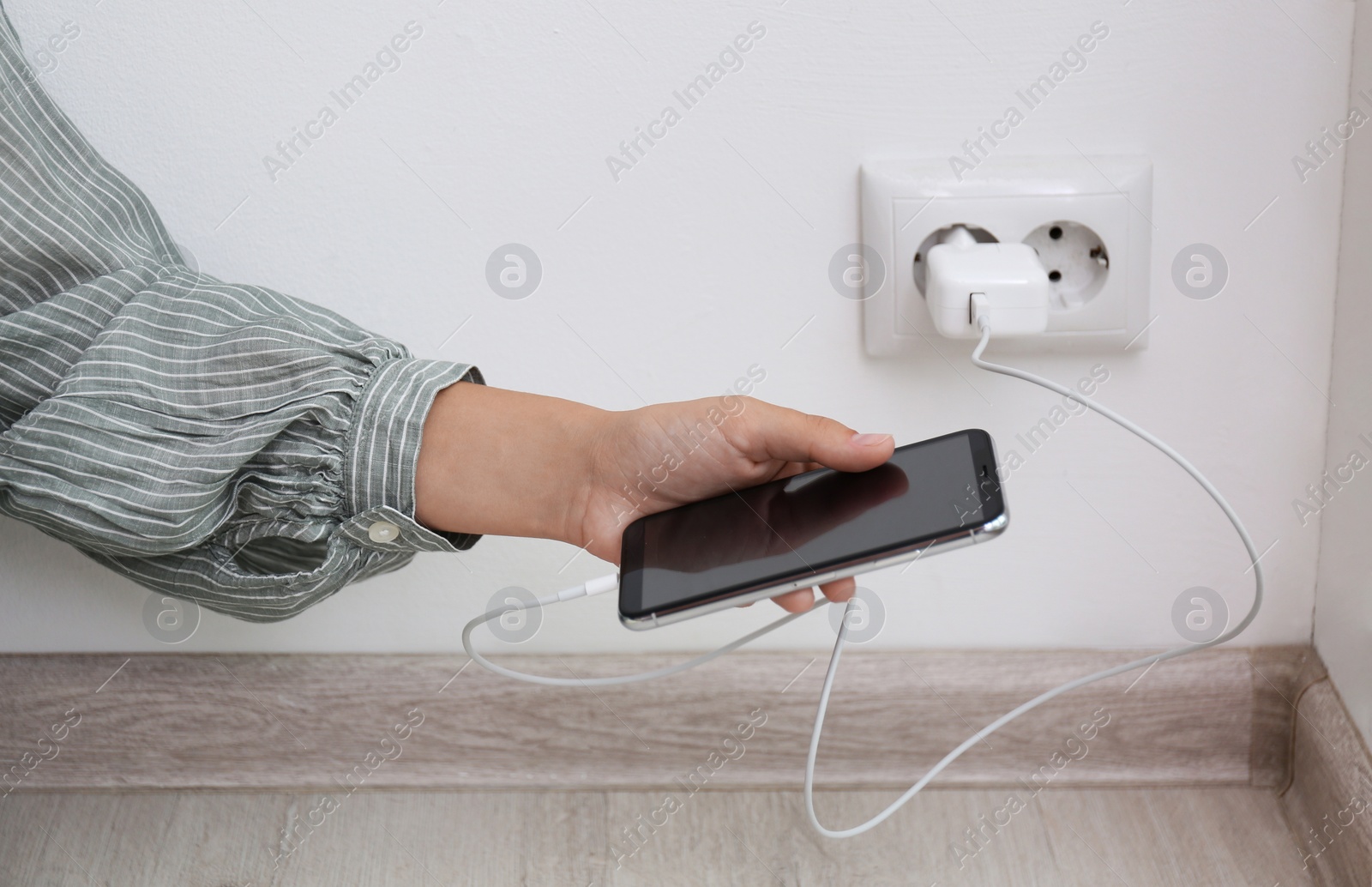 Photo of Woman holding mobile phone with connected charger near white brick wall indoors, closeup